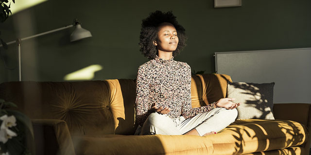 Eine junge Frau sitzt im Schneidersitz auf einem Sofa in ihrem Wohnzimmer - Sonnenstrahlen scheinen auf die Szene