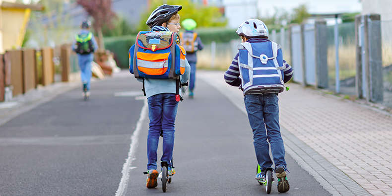 Zwei Kinder auf dem Roller mit Schulranzen