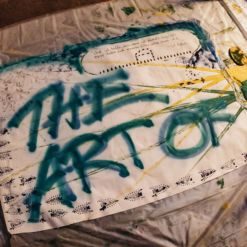 Ein Foto einer Malfläche, die mit dem gesprühten Schriftzug "the art of" und kreativen Linien spritzern versehen ist. Der weiße Stoff liegt auf dem Boden, daneben er erkennen einen Farbeimer.