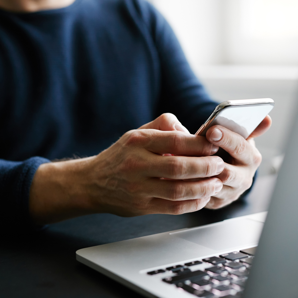 Mann sitzt am Laptop und sucht auf seinem Smartphone Informationen zum Schutzbrief der Öffentlichen