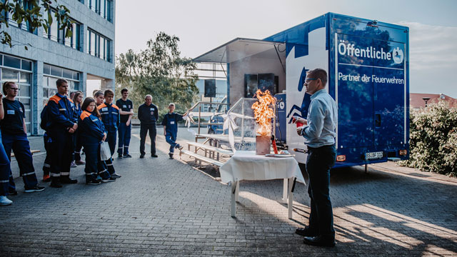Viele Menschen stehen vor dem blauen Kastenwagen, es wird auf einem Tisch ein Brand simuliert
