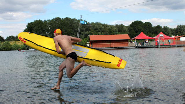 Junger Mann mit einem Schwimmbrett in gelb und dem DLRG-Logo rennt in ein Gewässer.