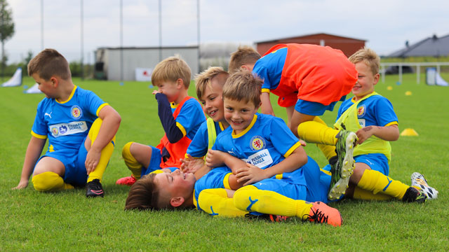 Mehrere Kinder in blau-gelben Fußballtrikots spielen auf dem Rasen, sie sehen aus als würden sie balgen.