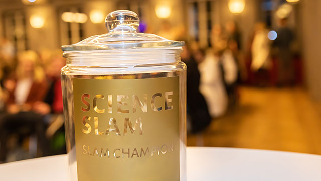 Ein Bild mit starkem Fokus auf einen Glasbehälter mit Deckel, auf dem Science Slam steht in Gold. Es ist die Trophäe des gleichnamigen Wettbewerbs.