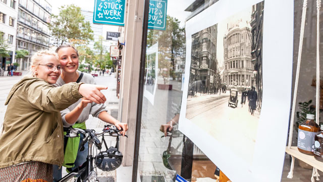 Copyright Andreas Rudolph; das Bild zeigt zwei junge Frauen, die ein Fahrrad schieben und vor einem Schaufenster stehen. Eine Person zeigt auf das augehängte Foto, eine alte Ansicht der Kultviertel-Straße