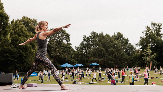 Eine Menge Menschen machen unter freiem Himmel Yoga, im Vordergrund steht eine Yogalehrerin in einer Krieger-Pose