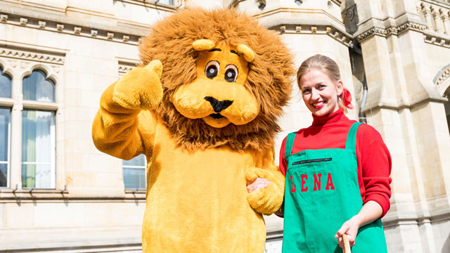 Lena und Leo vor dem Rathaus. Ein Mensch in einem Löwenkostüm steht zusammen mit einer Frau in einer grünen Schürze vor einem Gebäude. Der Löwe macht die Geste Daumen nach oben.