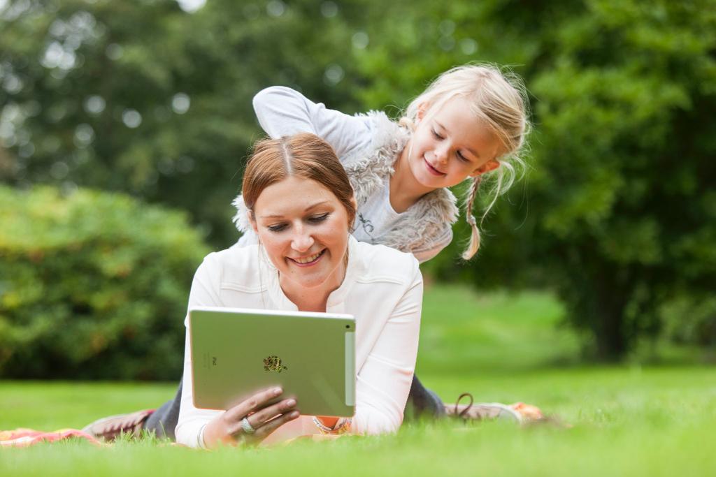 Junge Frau liegt gemeinsam mit ihrer Tochter auf einer Wiese und liest auf einem Tablet, das Kind klettert auf den Rücken der Mutter.