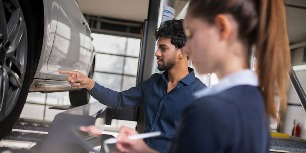 Im Schaden-Schnell-Service. Zwei Personen stehen neben einem Auto, von dem nur ein kleiner Teil zu sehen ist, auf einer Hubbühne. Die Person im hinteren Teil des Bildes ist ein Mann mit Bart, er zeigt auf das Auto. Im Vordergrund macht sich eine FRau mit einem Zopf Notizen dazu.