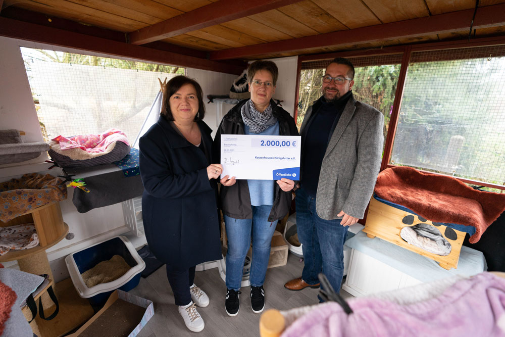 Drei Personen stehen in einem Raum, der für die Katzen hergerichtet wurde, sie zeigen einen Spendencheck der Öffentlichen mit der Summe von 2.000 Euro in die Kamera.