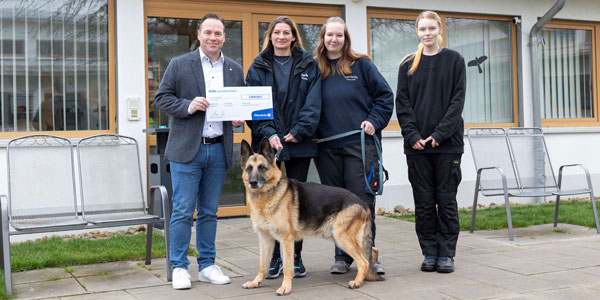 Vier Menschen stehen vor einem Haus und halten einen großen Scheck in die Kamera. Ein sehr netter Schäferhund steht vor den Menschen.