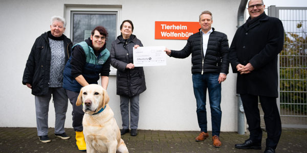 Eine Gruppe Menschen steht vor einem Gebäude - es wird ein sehr großer Check überreicht, im Vordergrund sitzt ein heller Labrador und guckt lieb in die Kamera