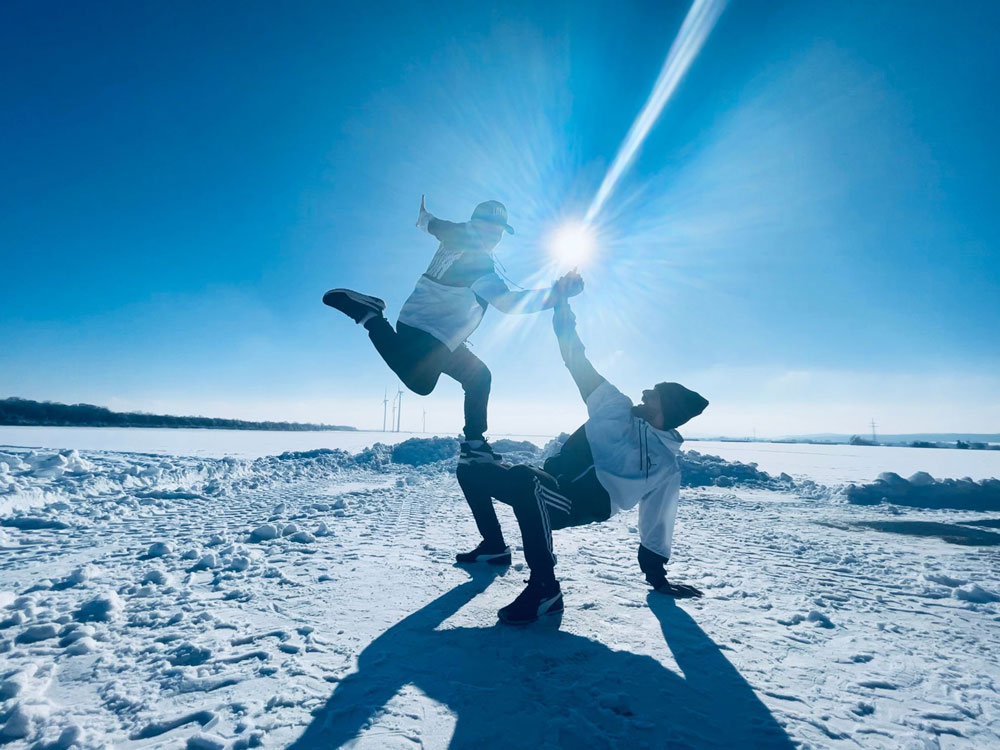 Zwei Tänzer in Winterjacken und Winterhosen im Schnee vor einem blauen und strahlendem Himmel. Es ist eine Hebefigur zu sehen, bei dem einer der beiden im Sprung von dem anderen gestützt wird