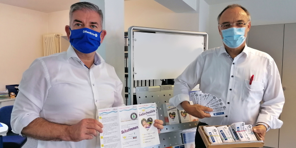 Zwei Mitarbeiter der Öffentlichen Versicherung tragen Mundnasenschutz und zeigen die Freikarten zum Verschenken