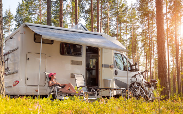 Ein Campingfahrzeug steht im Wald. Davor ist eine Frau, die sich auf einem Stuhl unter der ausgefahrenen Markise des Fahrzeugs ausruht. Ein paar Fahrräder stehen ebenfalls vor dem Wohnmobil.