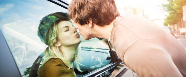 Eine Frau und ein Mann küssen sich, die Frau sitzt im Auto, der Mann steht vor dem Seitenfenster