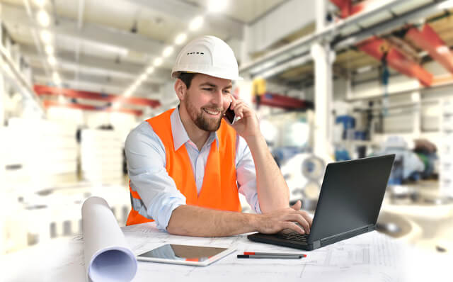 Mann in orangener Weste und weißen Helm, telefoniert und arbeitet am Laptop