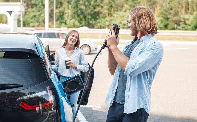 Ein Paar scherzt miteinander. neben ihnen steht ihr Auto an einer Elektrotankstelle